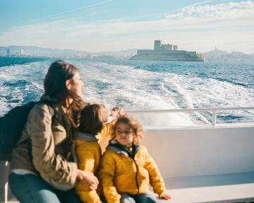 Una mujer y dos niños, con chaquetas amarillas, están sentados en un barco en el mar, frente al Castillo de If frente a Marsella, con la estela extendiéndose detrás de ellos.