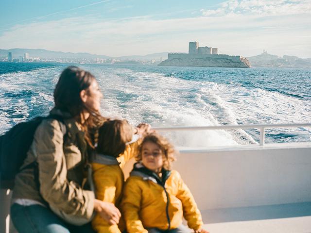 Une femme et deux enfants, vêtus de vestes jaunes, sont assis sur un bateau en mer, face au Château d’If au large de Marseille, avec le sillage qui se déploie derrière eux.